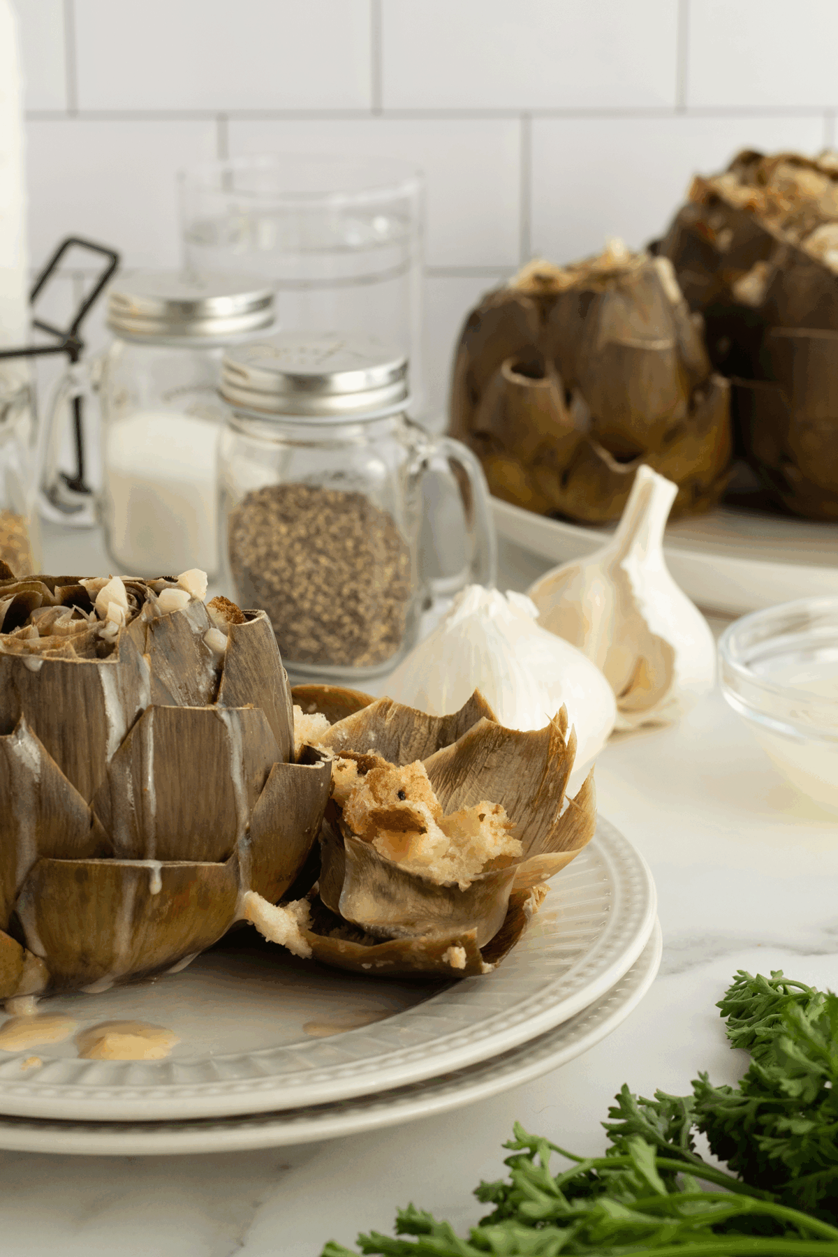 A plate with stuffed artichokes sits on a kitchen counter next to a whole garlic bulb, a glass bowl, fresh parsley, and jars of sugar and pepper. More stuffed artichokes and ingredients are in the background.