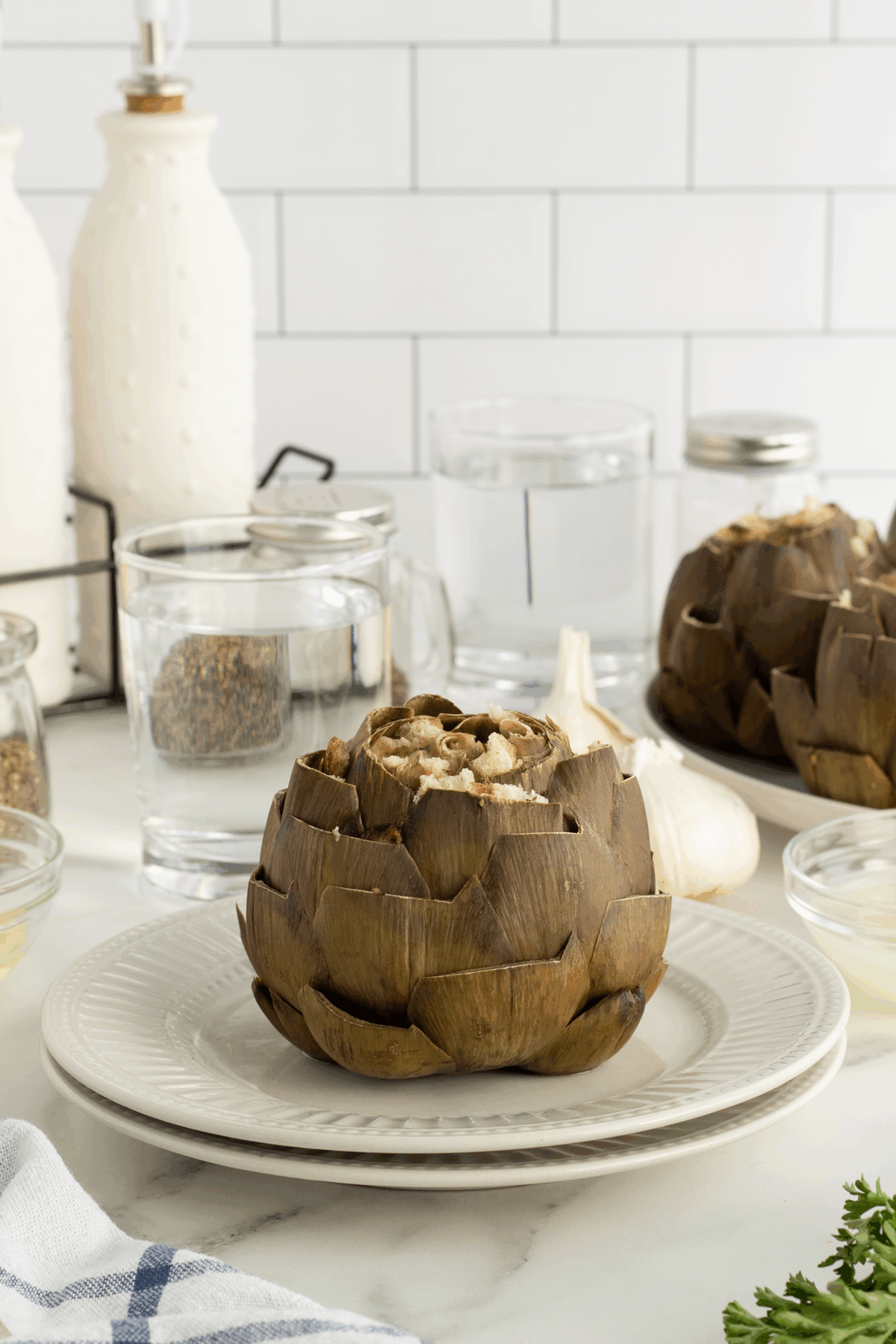 A cooked artichoke sits on a white plate with a glass of water and jars in the background, set on a white counter with garlic and a folded napkin nearby.