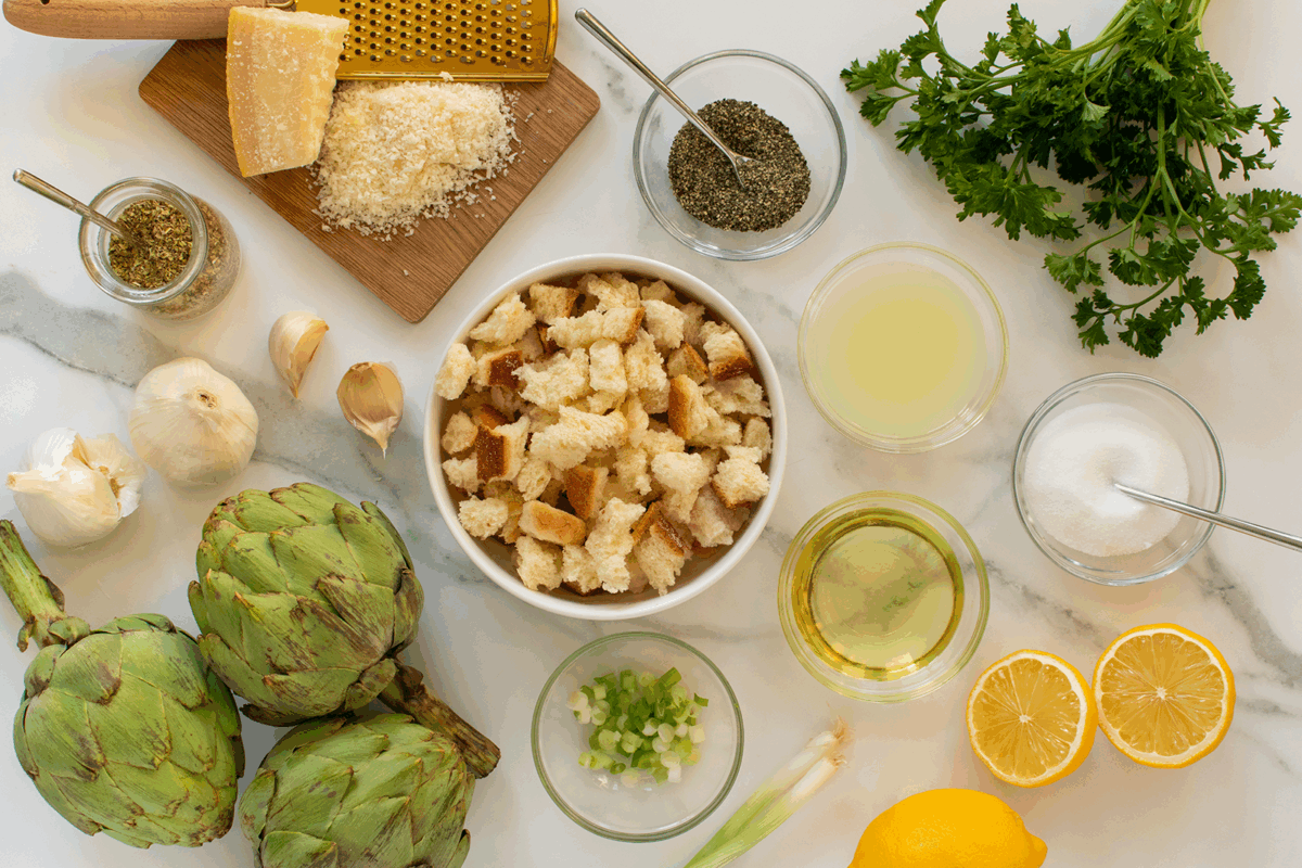 Overhead view of ingredients on a white surface, including artichokes, bread cubes, lemon halves, parsley, grated cheese, garlic, olive oil, salt, pepper, oregano, green onions, and lemon juice.