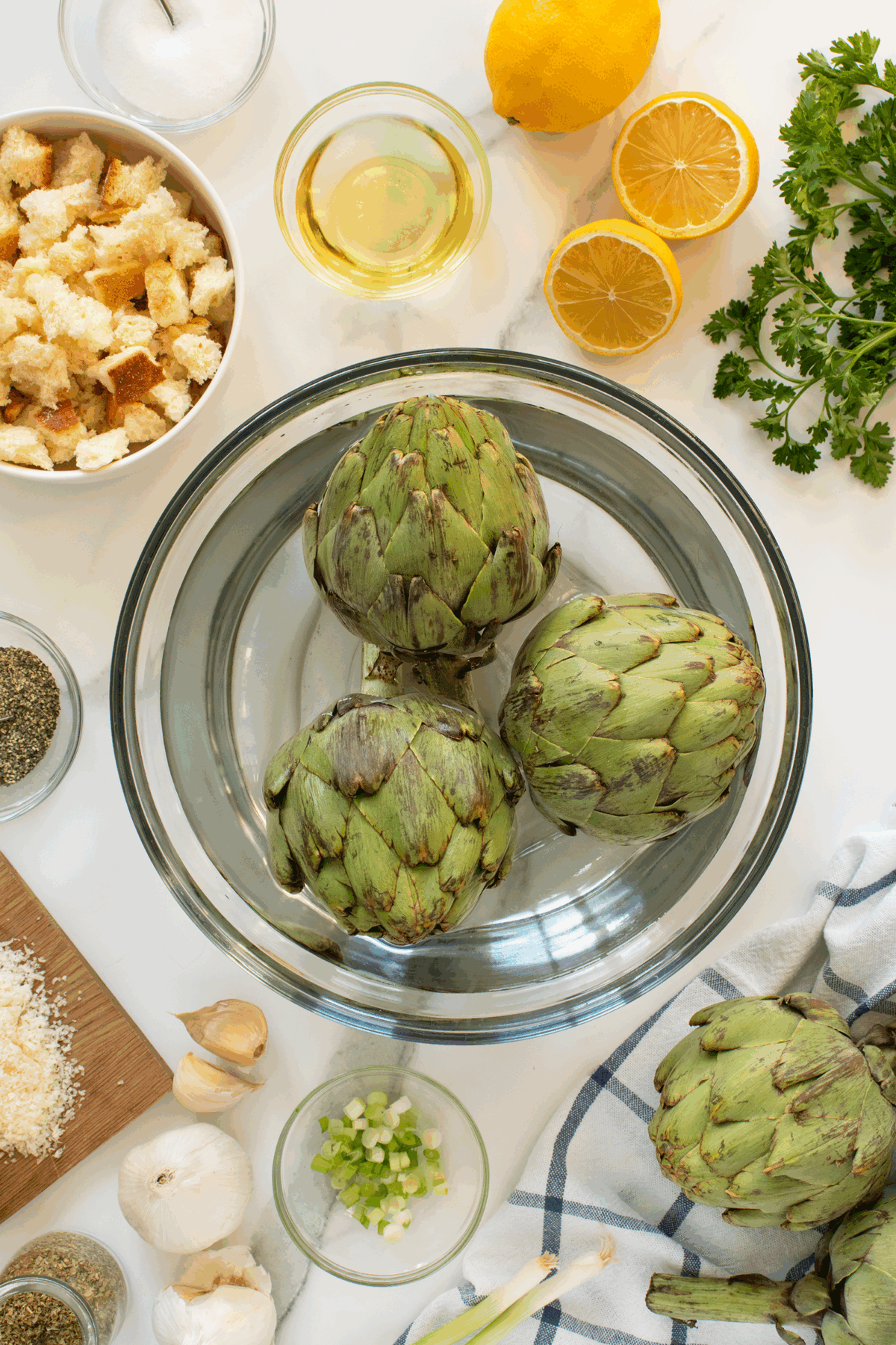 Overhead view of three fresh artichokes in a glass bowl, surrounded by ingredients such as lemon halves, parsley, cubed bread, oil, grated cheese, garlic, green onions, pepper, and a striped towel on a white surface.