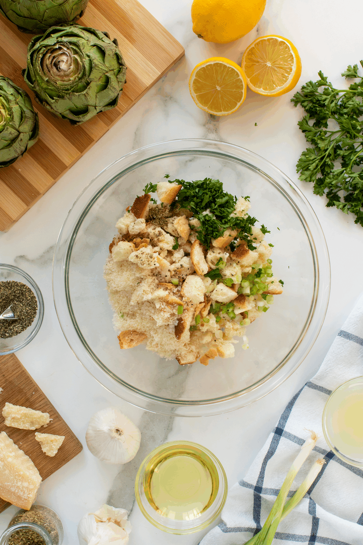 A glass bowl containing chopped bread, parsley, cheese, and seasonings sits on a marble counter surrounded by fresh artichokes, lemon halves, parsley, olive oil, garlic, black pepper, and a kitchen towel.