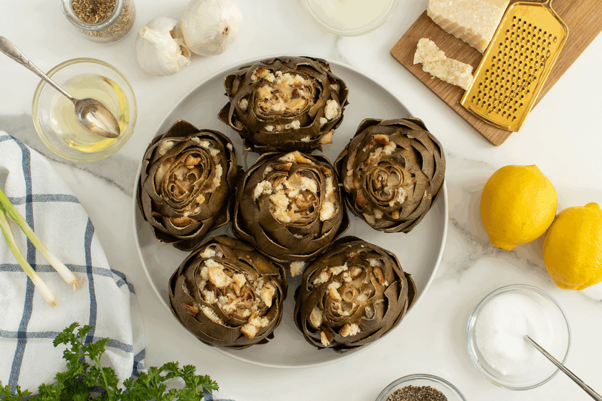 A white plate with five stuffed artichokes is on a marble surface, surrounded by garlic, lemons, grated cheese, parsley, a striped towel, spices, and bowls with ingredients.