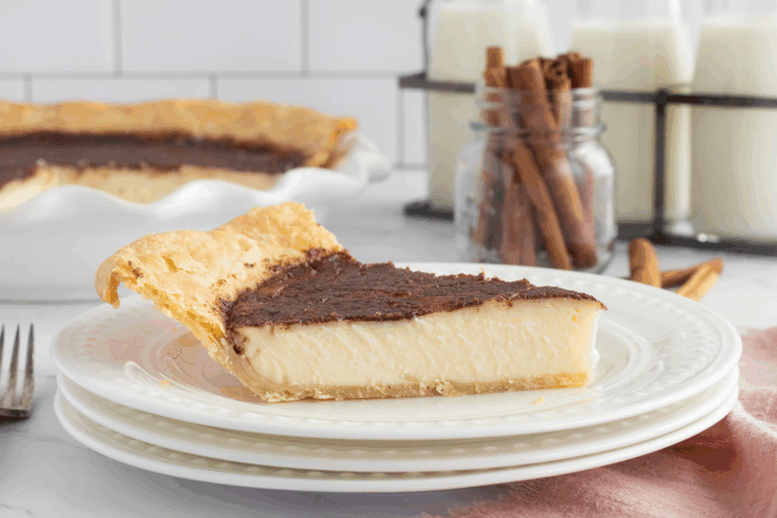 A slice of custard pie with a chocolate layer on top sits on a stack of white plates. In the background are the remaining pie, a jar of cinnamon sticks, and glasses of milk.