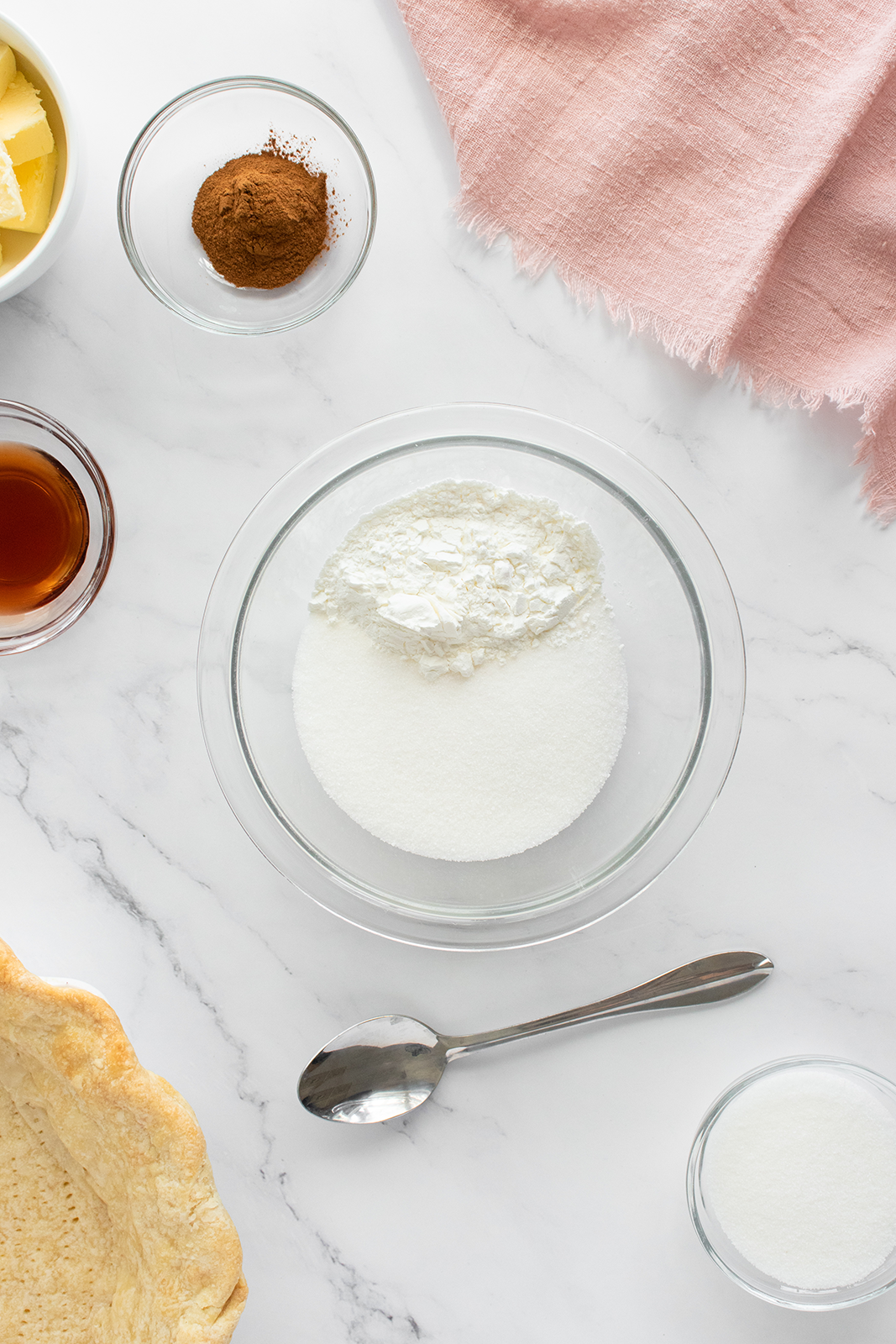 A glass bowl with white sugar and flour on a marble surface, surrounded by a spoon, a pie crust, a small bowl of cinnamon, a cup of syrup, a dish of butter, a glass of sugar, and a pink cloth.