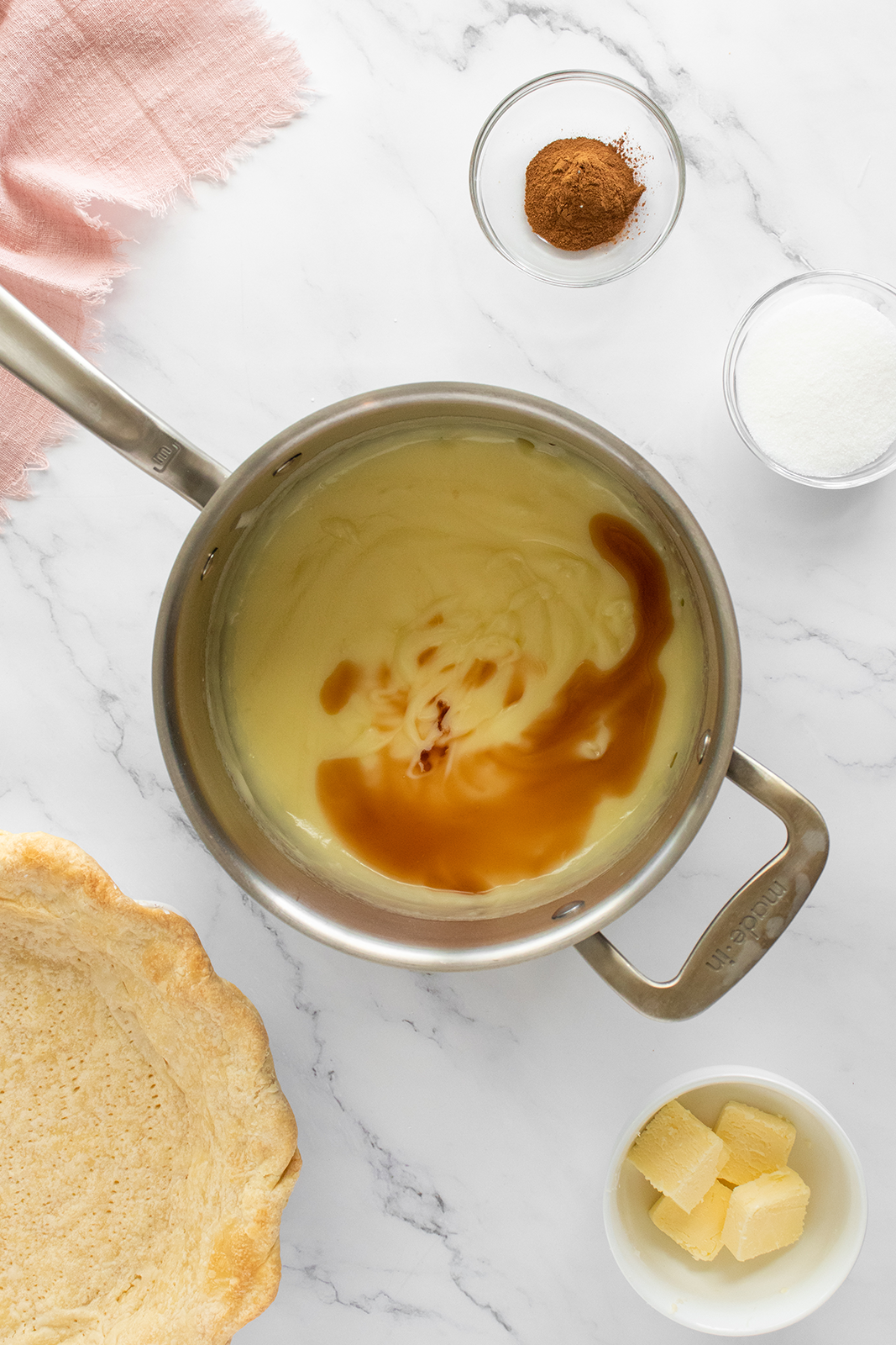 A saucepan with custard and vanilla extract on a marble surface, surrounded by a baked pie crust, cubes of butter, a bowl of sugar, and a bowl of cinnamon. A pink cloth is partially visible in the top left corner.