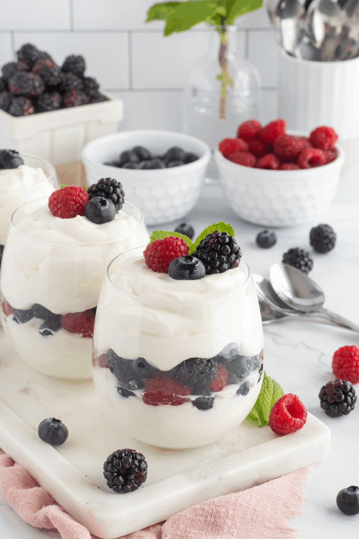 Two glasses filled with layered creamy yogurt and fresh berries—blackberries, raspberries, and blueberries—garnished with berries and mint. Bowls of berries and spoons are in the background on a white marble surface.