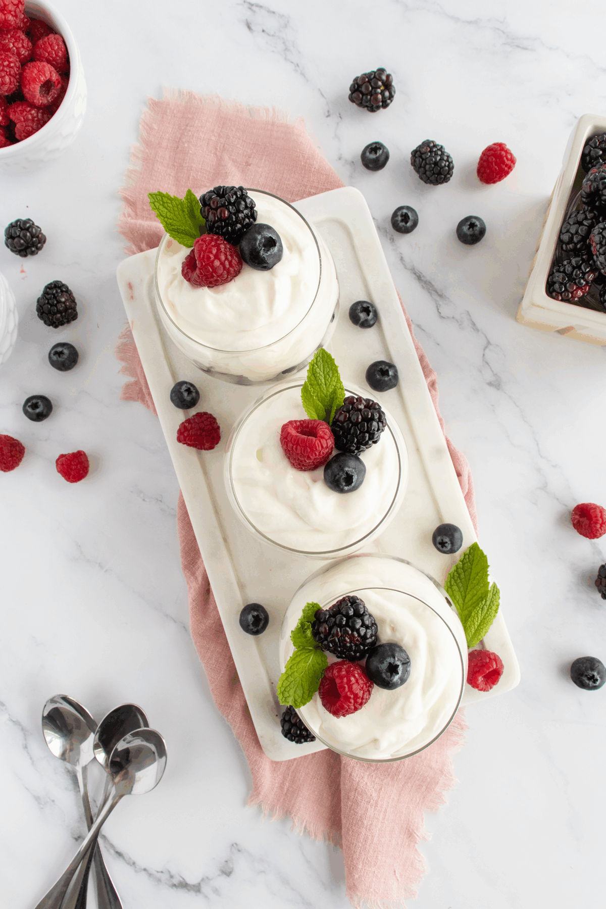 Three glass cups of creamy dessert topped with raspberries, blackberries, blueberries, and mint leaves are arranged on a tray over a pink cloth. Loose berries and spoons are scattered on a white marble surface.