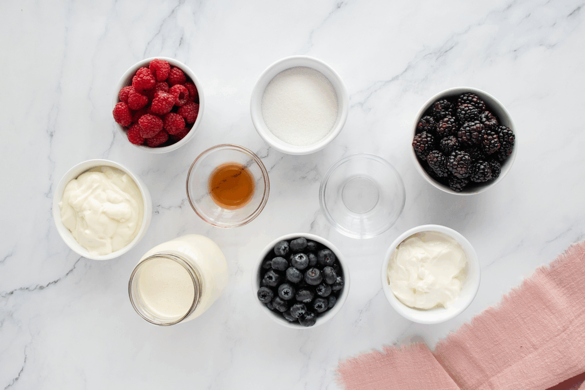 Overhead view of bowls and jars containing raspberries, blackberries, blueberries, sugar, yogurt, cream, vanilla extract, and an empty glass, arranged on a white marble surface with a pink cloth in the corner.