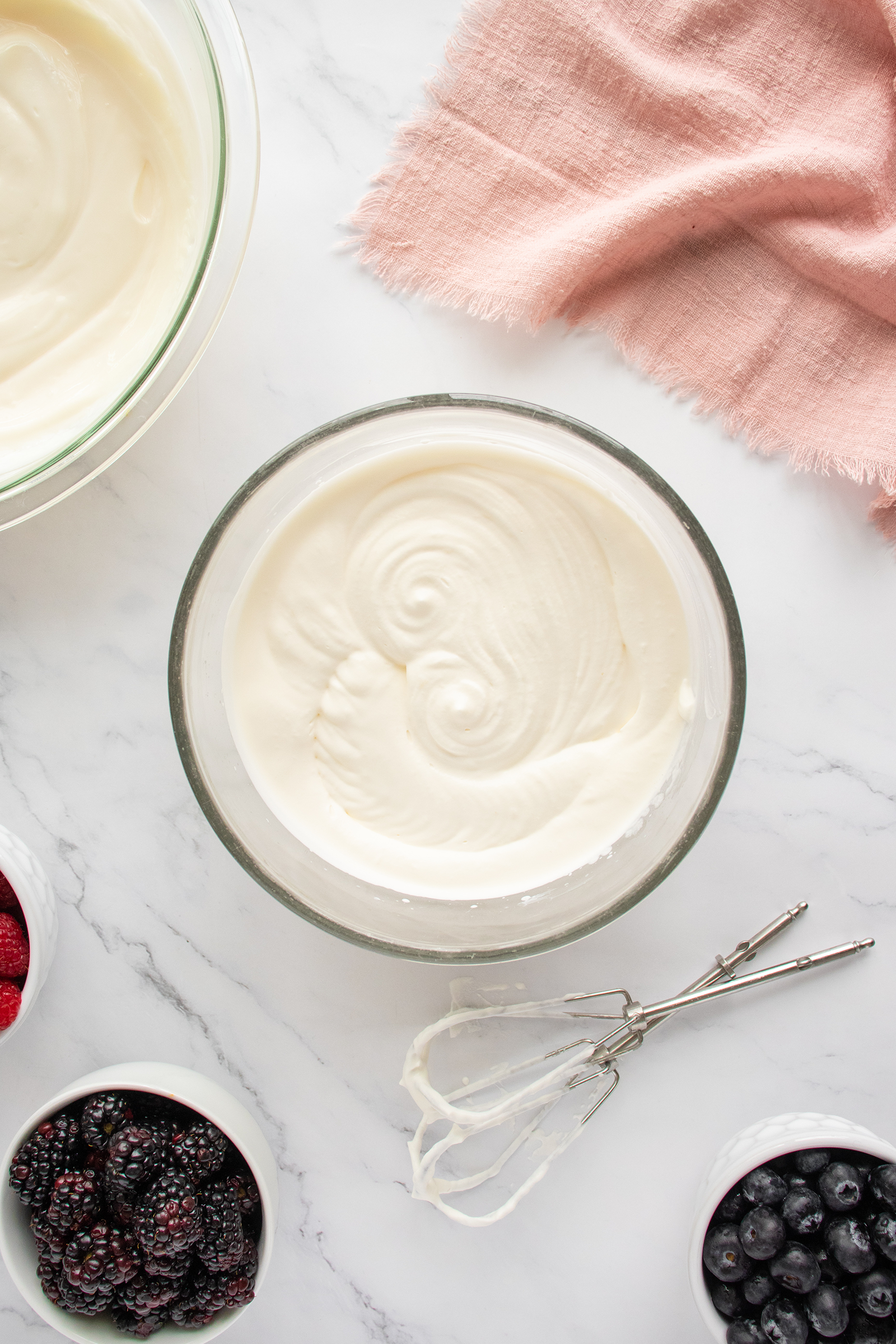 A bowl of creamy white mixture sits on a marble surface, surrounded by bowls of fresh berries, an electric mixer with beaters, and a pink cloth in the top right corner.