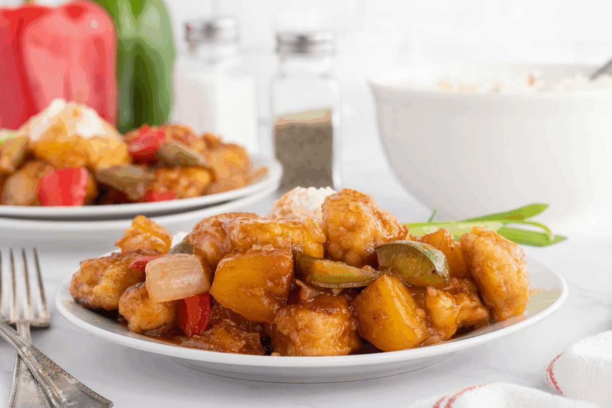 A plate of sweet and sour chicken with chunks of pineapple, bell peppers, and onions served with white rice. A bowl of rice and another plate of food are visible in the background.