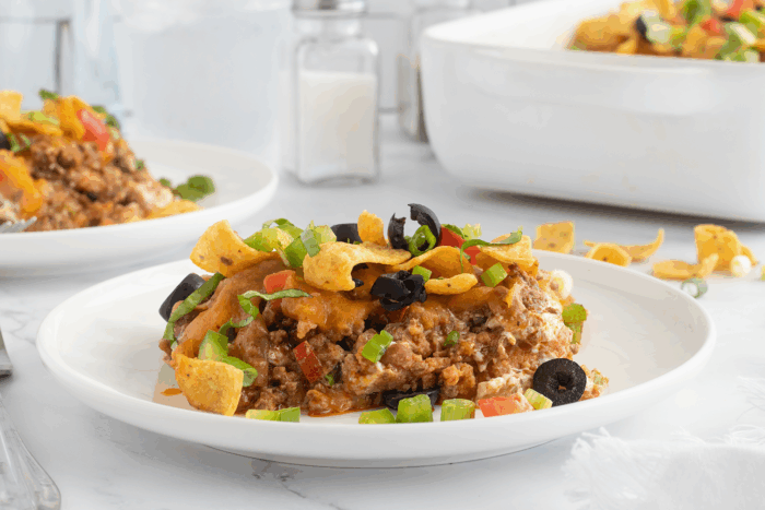 A serving of taco casserole topped with Fritos, sliced black olives, chopped green onions, and melted cheese on a white plate, with a casserole dish and condiments in the background.