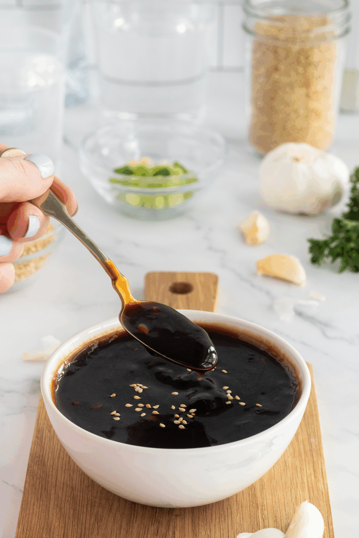 A hand holds a spoon over a bowl filled with dark sauce topped with sesame seeds. In the background are garlic cloves, green onions, and glass jars on a white countertop.