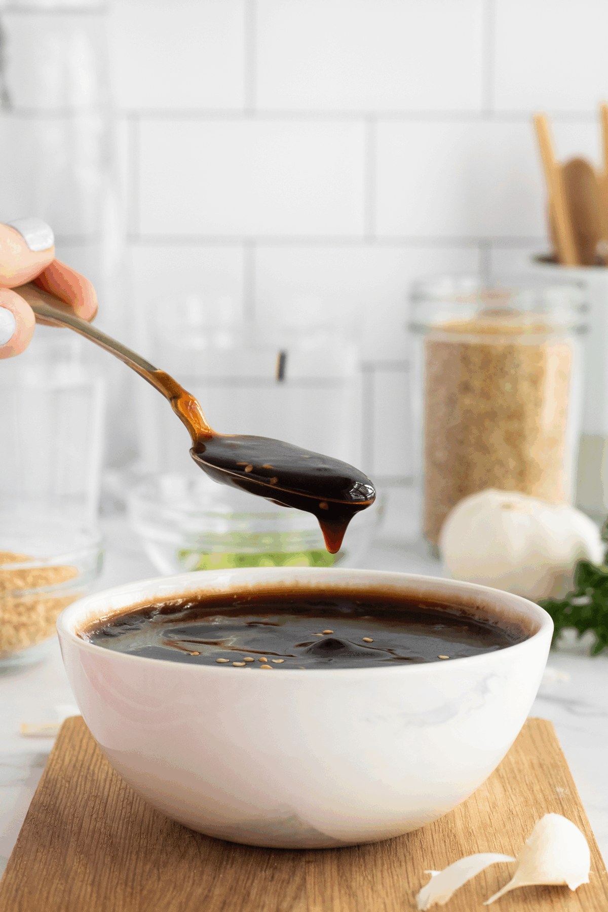 A hand holds a spoonful of thick, dark sauce above a white bowl filled with the same sauce. The bowl sits on a wooden surface; garlic, sesame seeds, and glass jars are in the background.