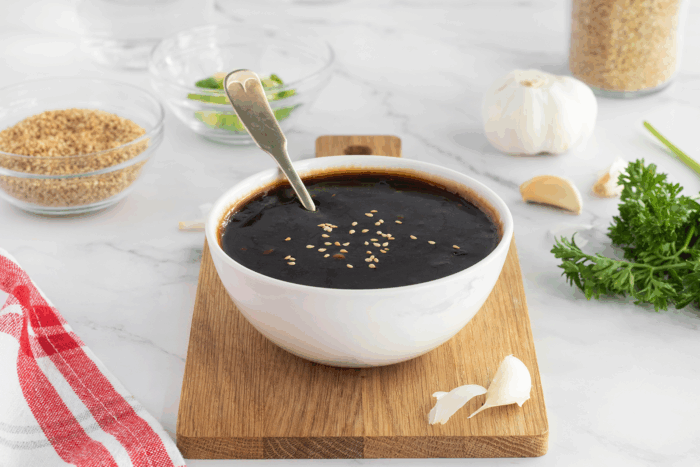 A white bowl filled with dark soy sauce, topped with sesame seeds and a spoon inside, sits on a wooden board. Garlic, parsley, sesame seeds, and chopped green onions are nearby on a white marble surface.