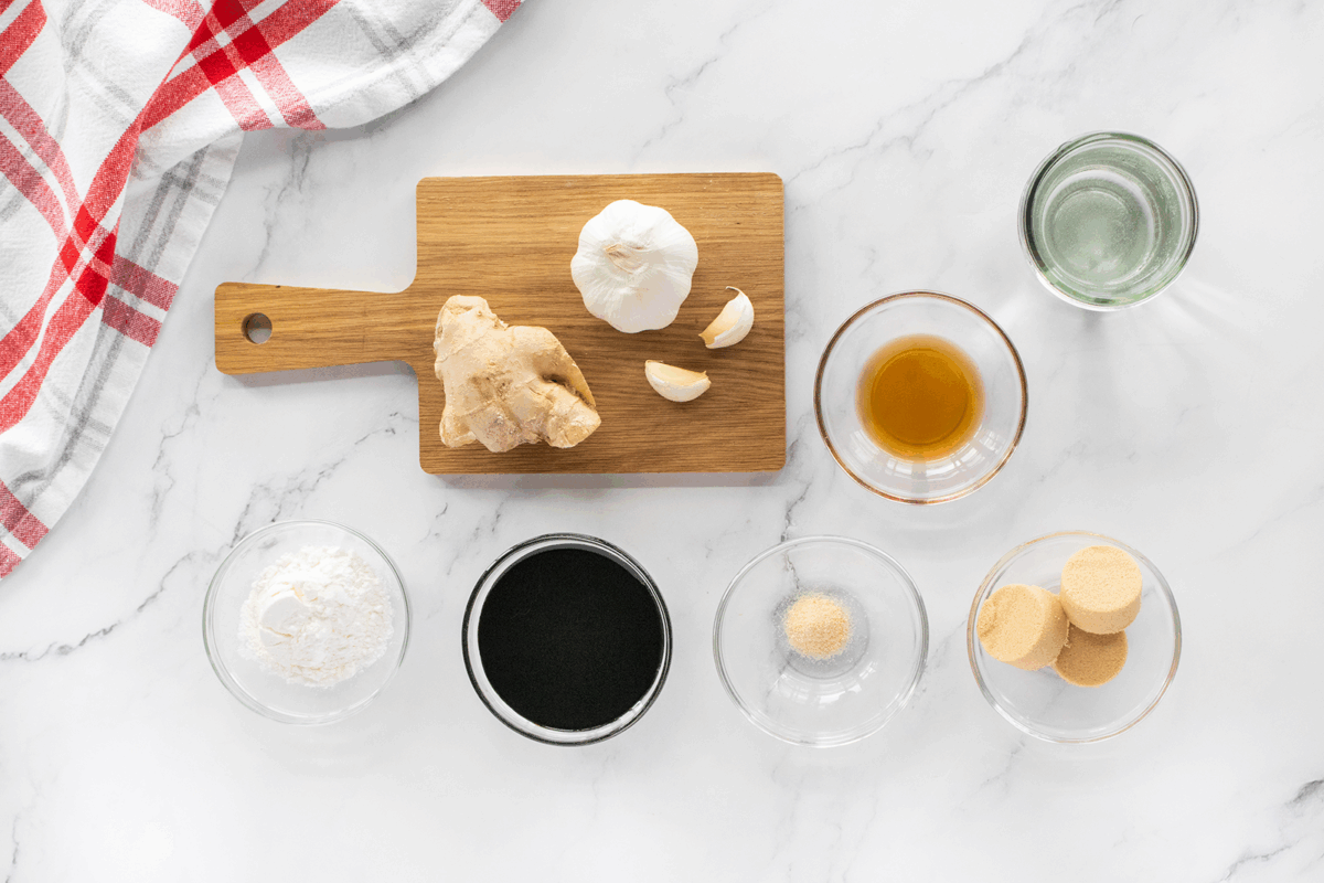 Ingredients for a recipe arranged on a marble surface: garlic bulb, ginger, small bowls of soy sauce, brown sugar, cornstarch, water, vinegar, and a red-and-white towel in the corner.