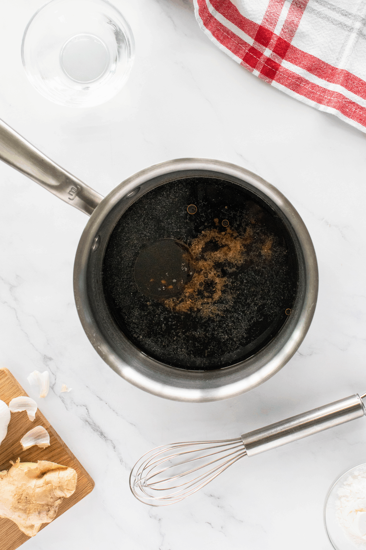 A saucepan filled with dark liquid on a white marble countertop, surrounded by a whisk, a cutting board with garlic and ginger, a towel with red stripes, and a glass bowl of water.