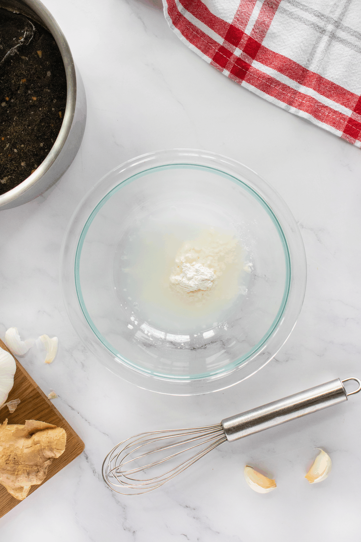 A glass bowl with white powder and liquid sits on a marble countertop near a whisk, garlic cloves, a piece of ginger on a cutting board, a metal pot, and a red-and-white kitchen towel.