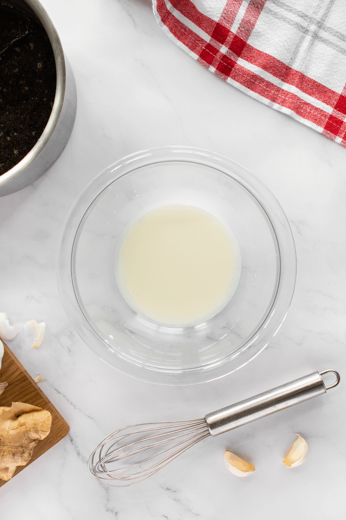 A glass bowl filled with a white liquid sits on a marble countertop, surrounded by a metal whisk, garlic cloves, ginger, a pan, and a red-and-white checkered kitchen towel.