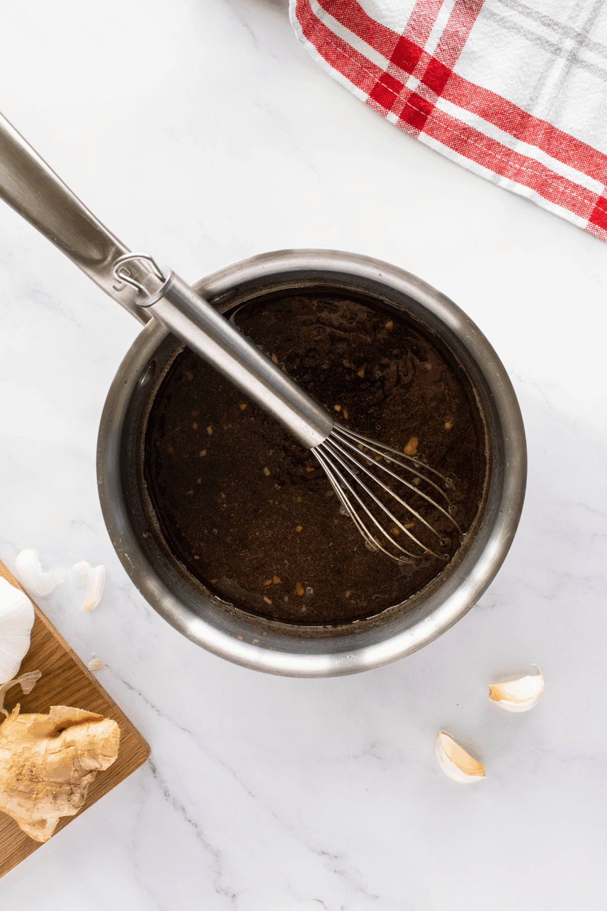 A metal bowl filled with dark sauce being whisked, surrounded by a red-and-white towel, garlic cloves, and a piece of ginger root on a white countertop.
