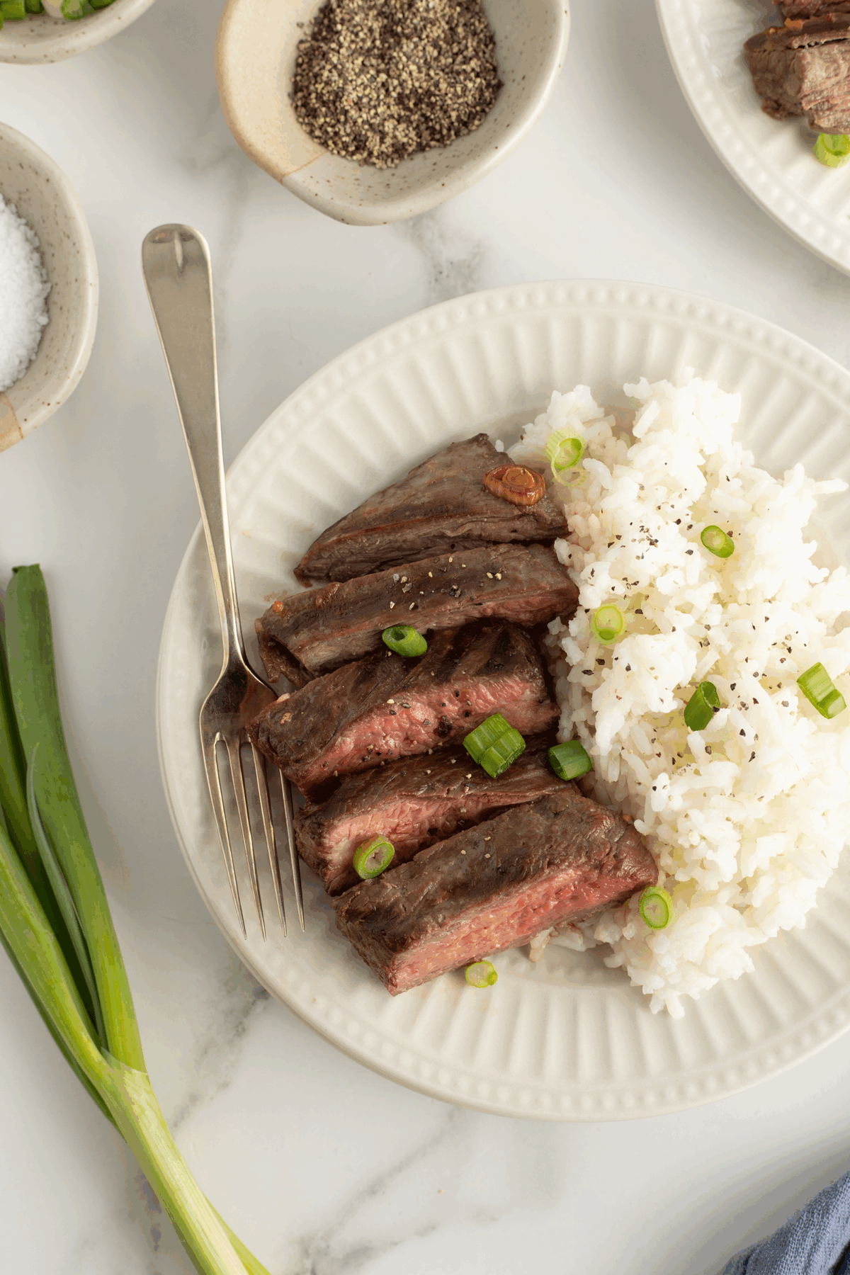 A white plate with sliced medium-rare steak and white rice, garnished with chopped green onions and black pepper. A fork rests on the plate, with salt, pepper, and a green onion beside it on a marble surface.