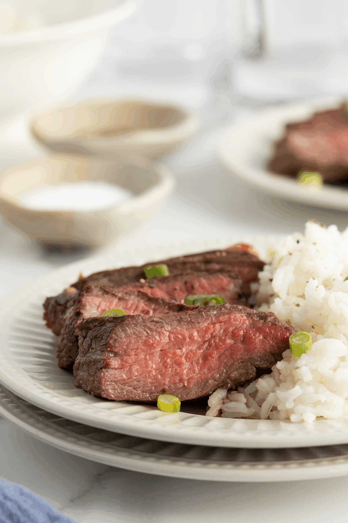 Slices of medium-rare steak garnished with green onions, served next to a portion of white rice on a white plate. Blurred bowls and another plate are visible in the background.