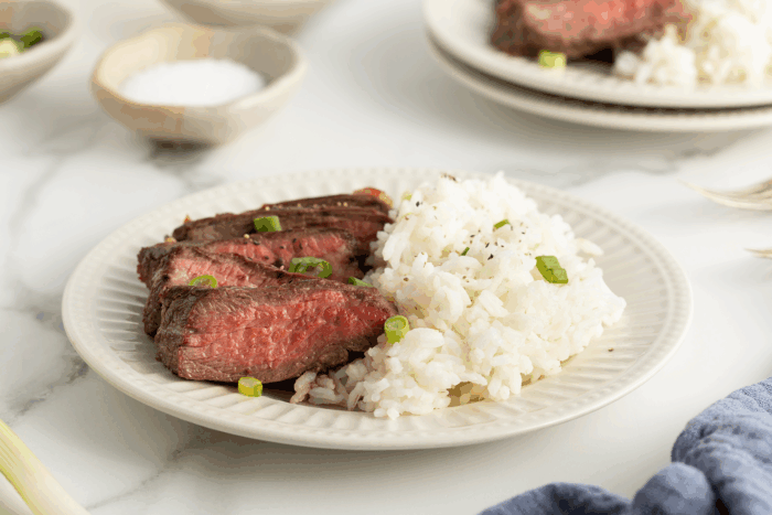A plate with sliced medium-rare steak and a serving of white rice, garnished with chopped green onions, sits on a white marble surface. Another plate of food and a bowl of salt are in the background.
