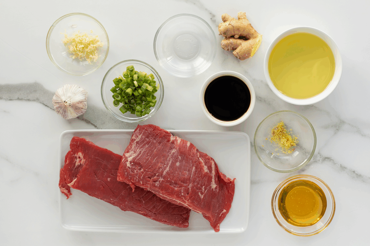 Two raw beef slices on a white tray are surrounded by bowls of chopped green onion, minced garlic, grated ginger, soy sauce, oil, honey, lemon zest, vinegar, and a whole garlic bulb on a marble countertop.