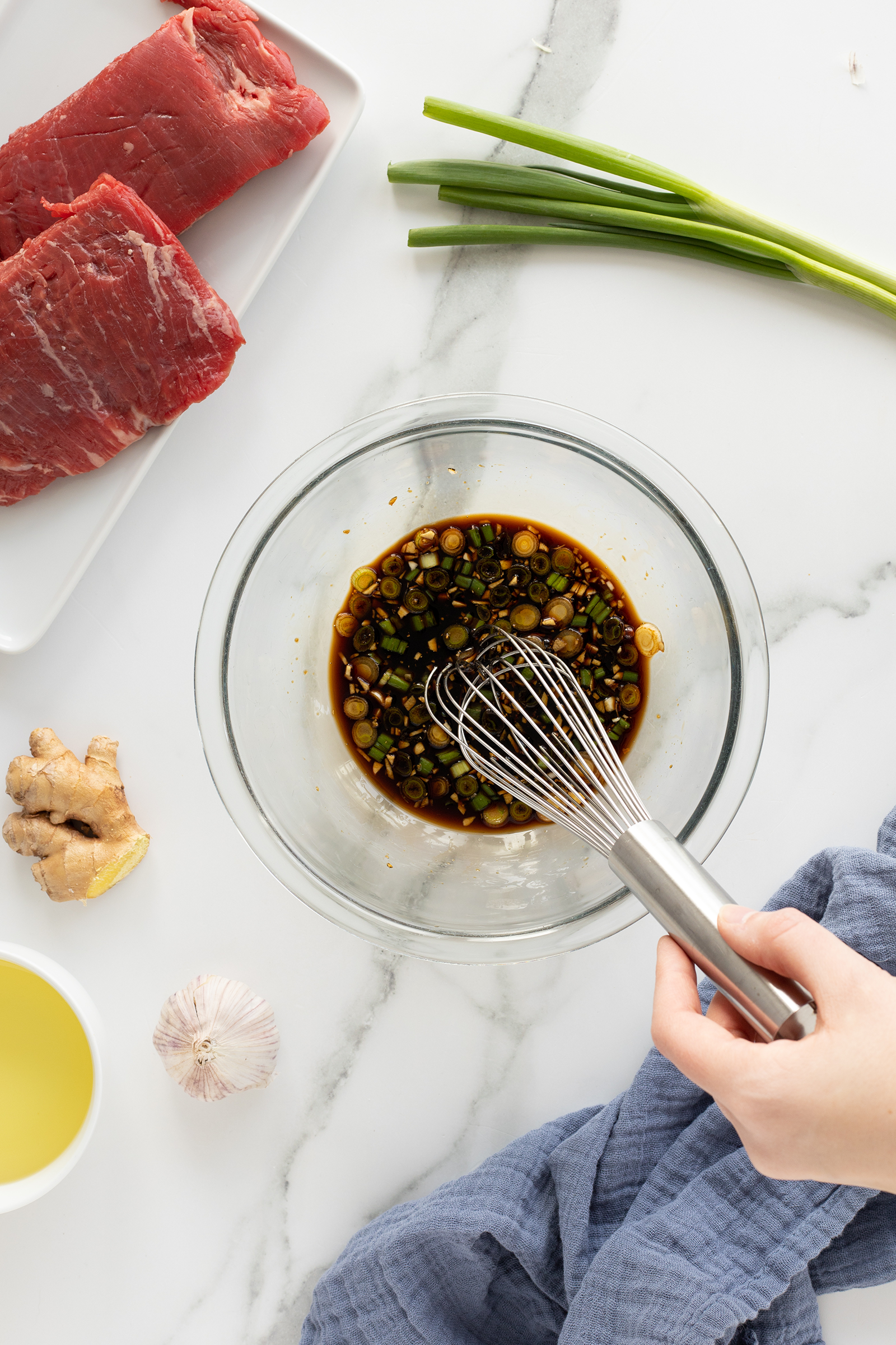 A hand whisks a soy-based marinade with chopped green onions in a glass bowl. Nearby are raw beef, green onions, ginger, garlic, and a cup of oil on a marble countertop.
