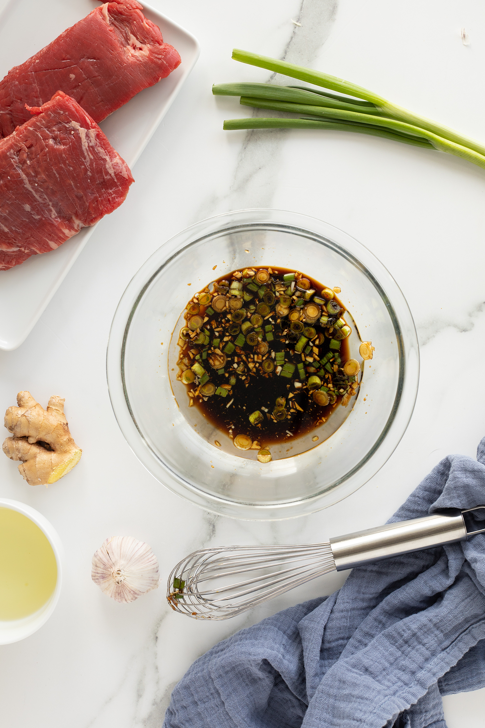 A bowl of soy sauce marinade with chopped green onions sits on a marble countertop, surrounded by raw beef, ginger, garlic, green onions, oil, and a whisk on a blue cloth.