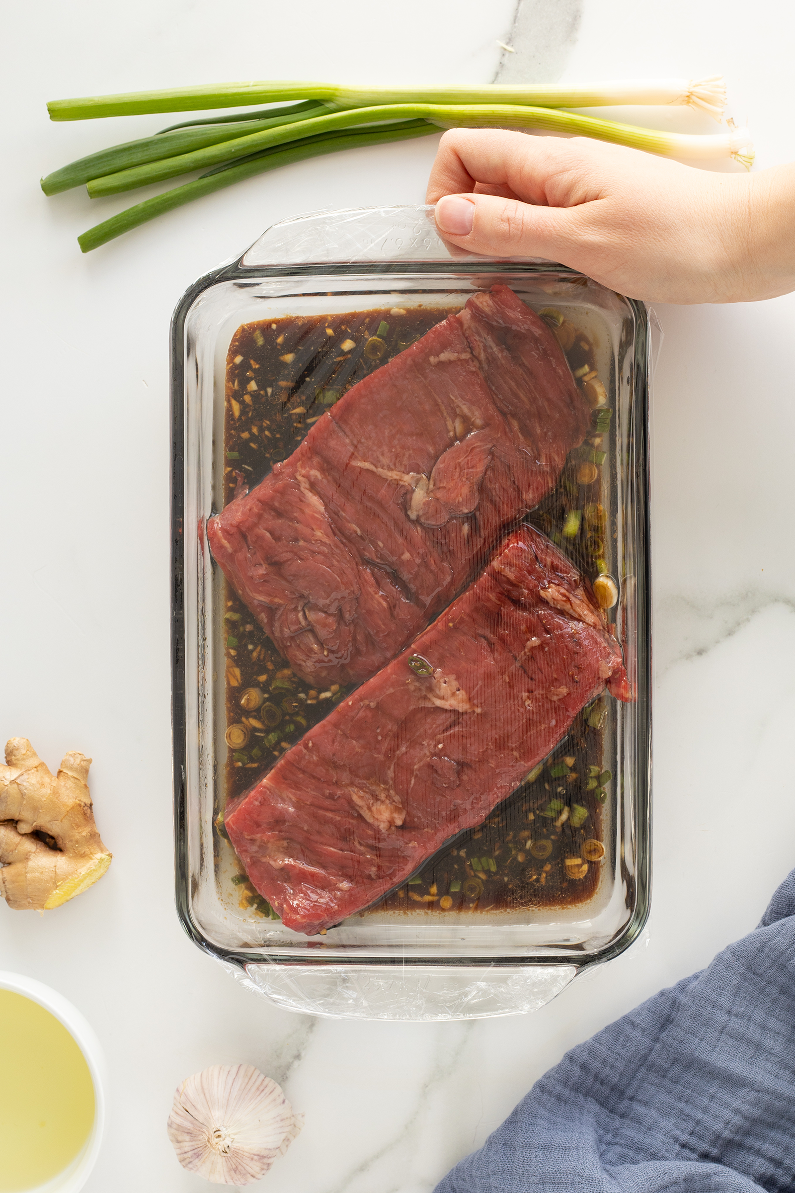 A hand covers a glass dish with two raw beef steaks marinating in a dark sauce. Green onions, ginger, garlic, and a bowl of oil are nearby on a white marble surface.