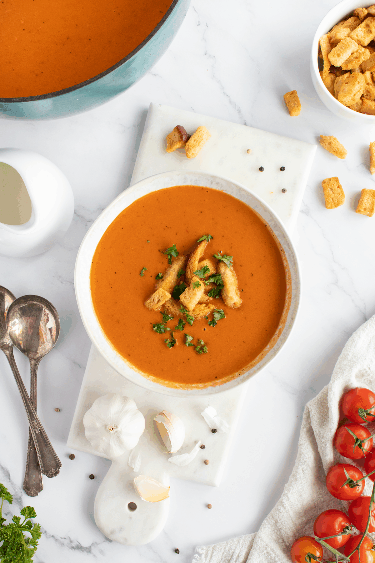 A bowl of tomato soup topped with croutons and parsley sits on a marble board, surrounded by garlic, tomatoes, croutons, a jug of cream, spoons, and peppercorns on a white surface.