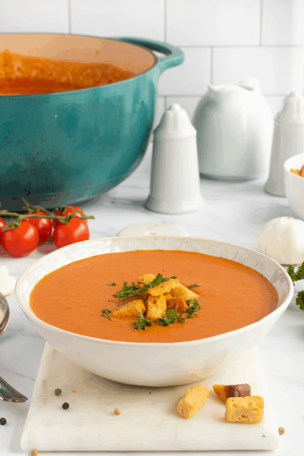 A bowl of creamy tomato soup topped with croutons and parsley sits on a marble board. In the background, theres a pot of soup, tomatoes on the vine, salt and pepper shakers, and a garlic bulb on a white counter.