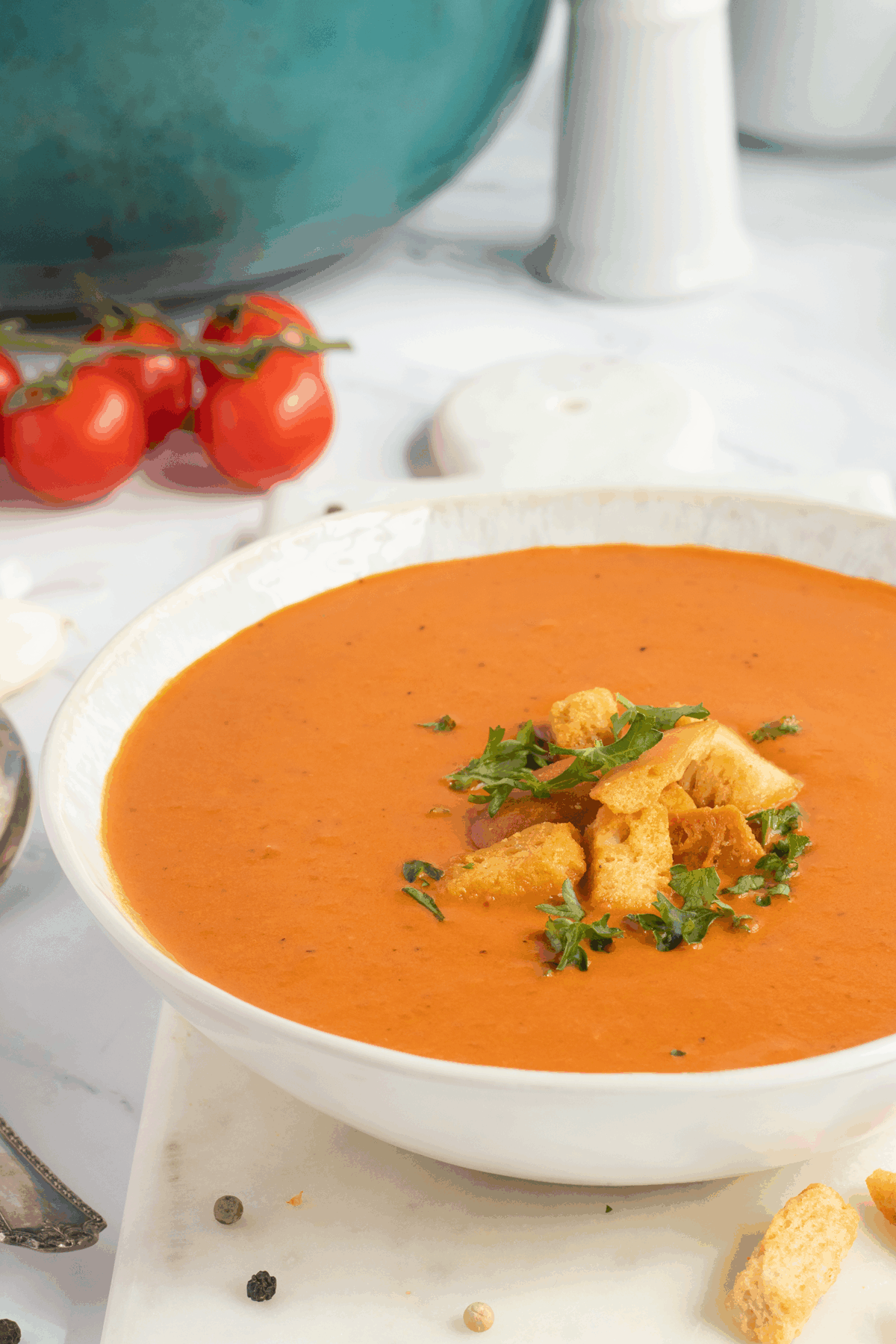 A bowl of creamy tomato soup topped with croutons and fresh herbs, set on a white surface with tomatoes, a pot, and a salt shaker in the background.