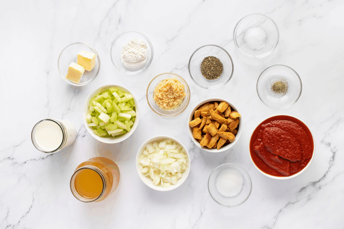 Various ingredients in small bowls on a white marble surface, including chopped celery, onions, croutons, tomato puree, butter, cream, broth, minced garlic, flour, salt, pepper, and sugar.