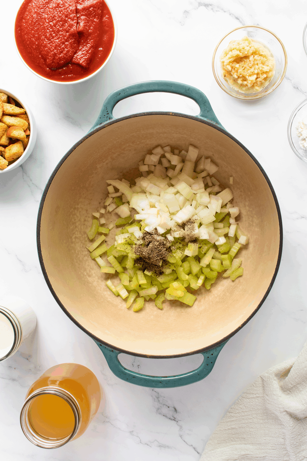 A large pot with chopped celery, onions, and seasonings inside, surrounded by bowls of tomato sauce, minced garlic, croutons, flour, milk, and broth on a white marble surface.