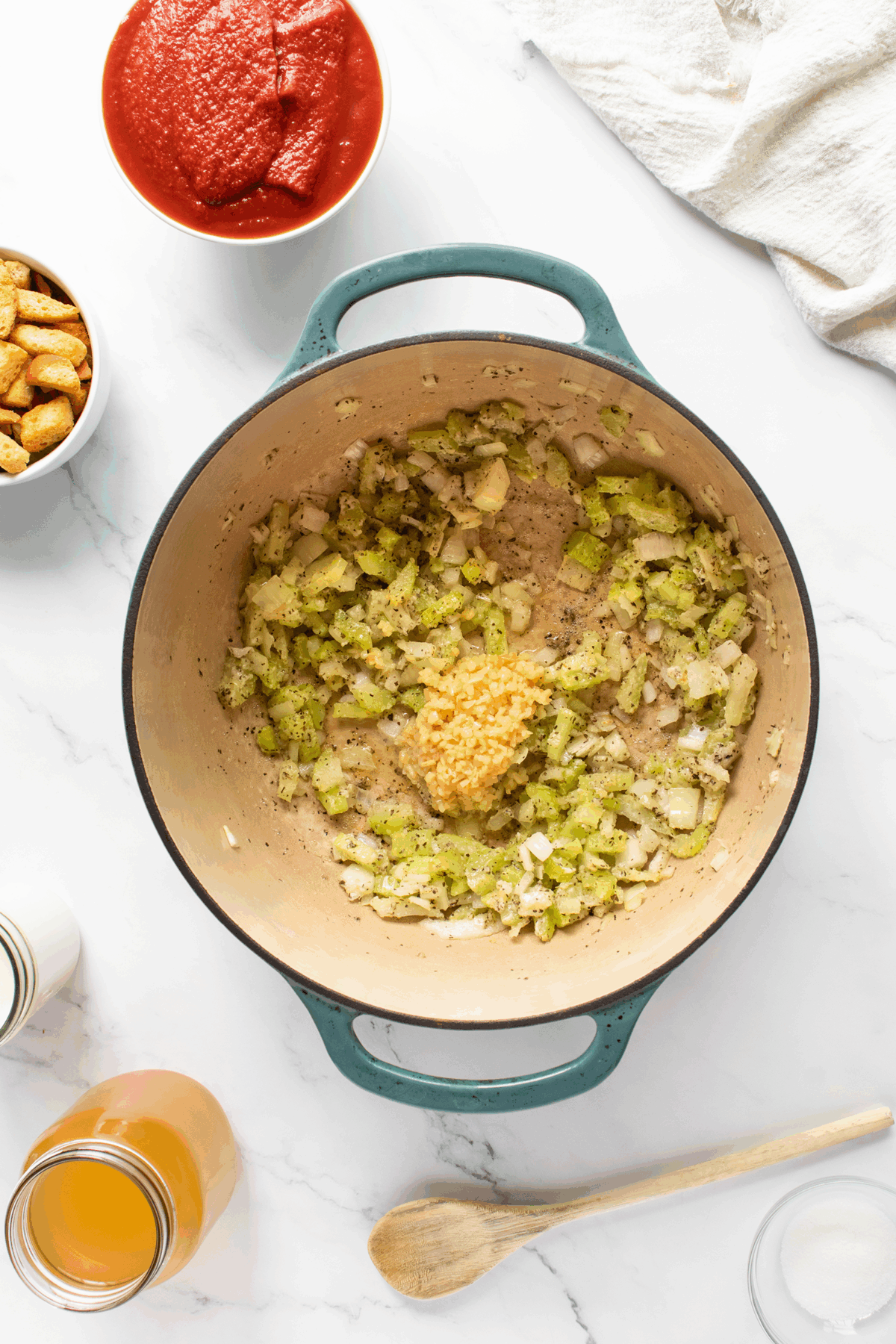 A pot filled with sautéed chopped onions, celery, and minced garlic sits on a white countertop. Surrounding it are bowls of tomato sauce, croutons, and jars of broth and cream, with a wooden spoon nearby.