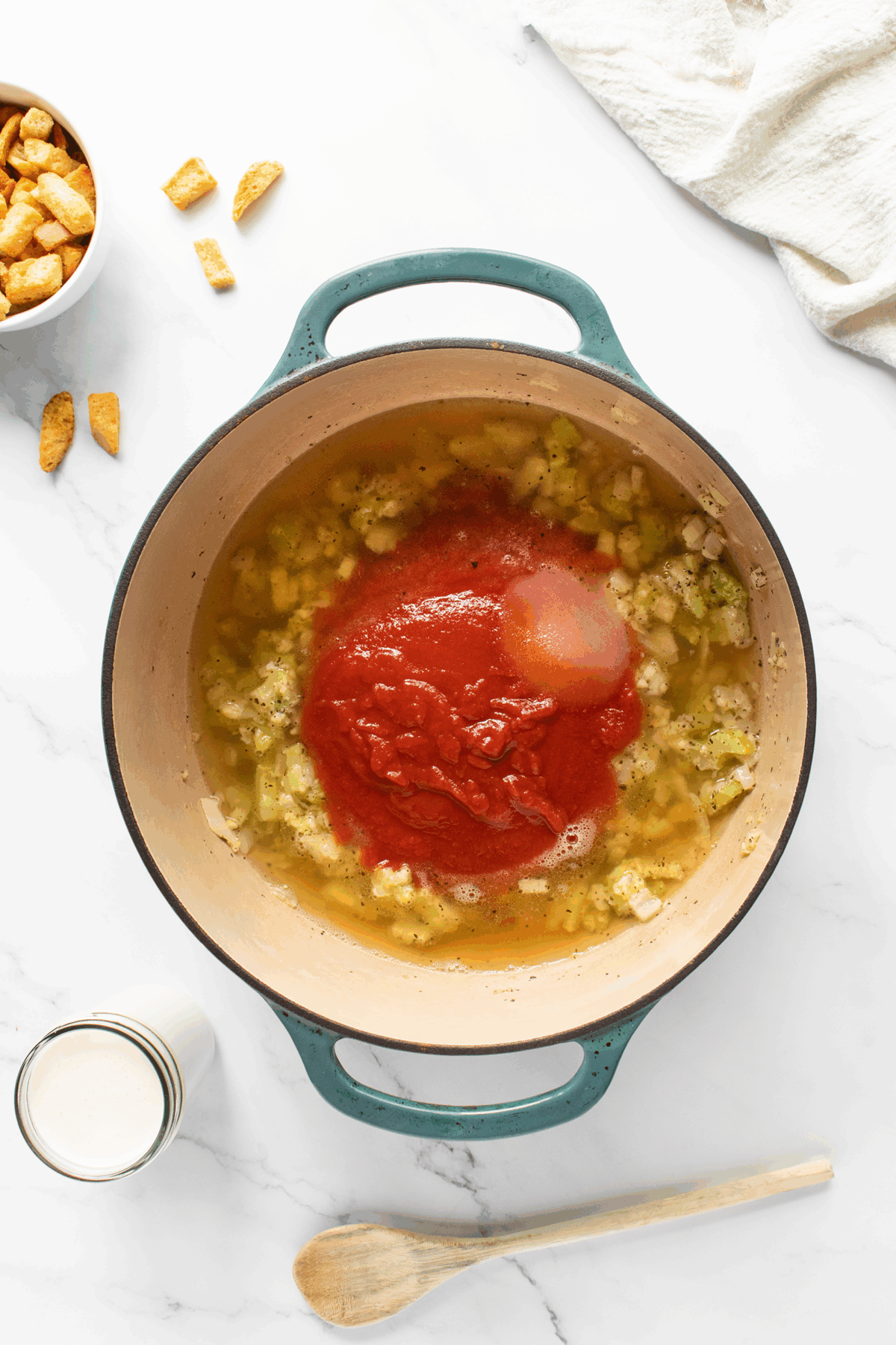 A pot on a white surface contains chopped onions and celery, broth, tomato sauce, and a mound of salt. Nearby are croutons, a jar of cream, a wooden spoon, and a folded white towel.