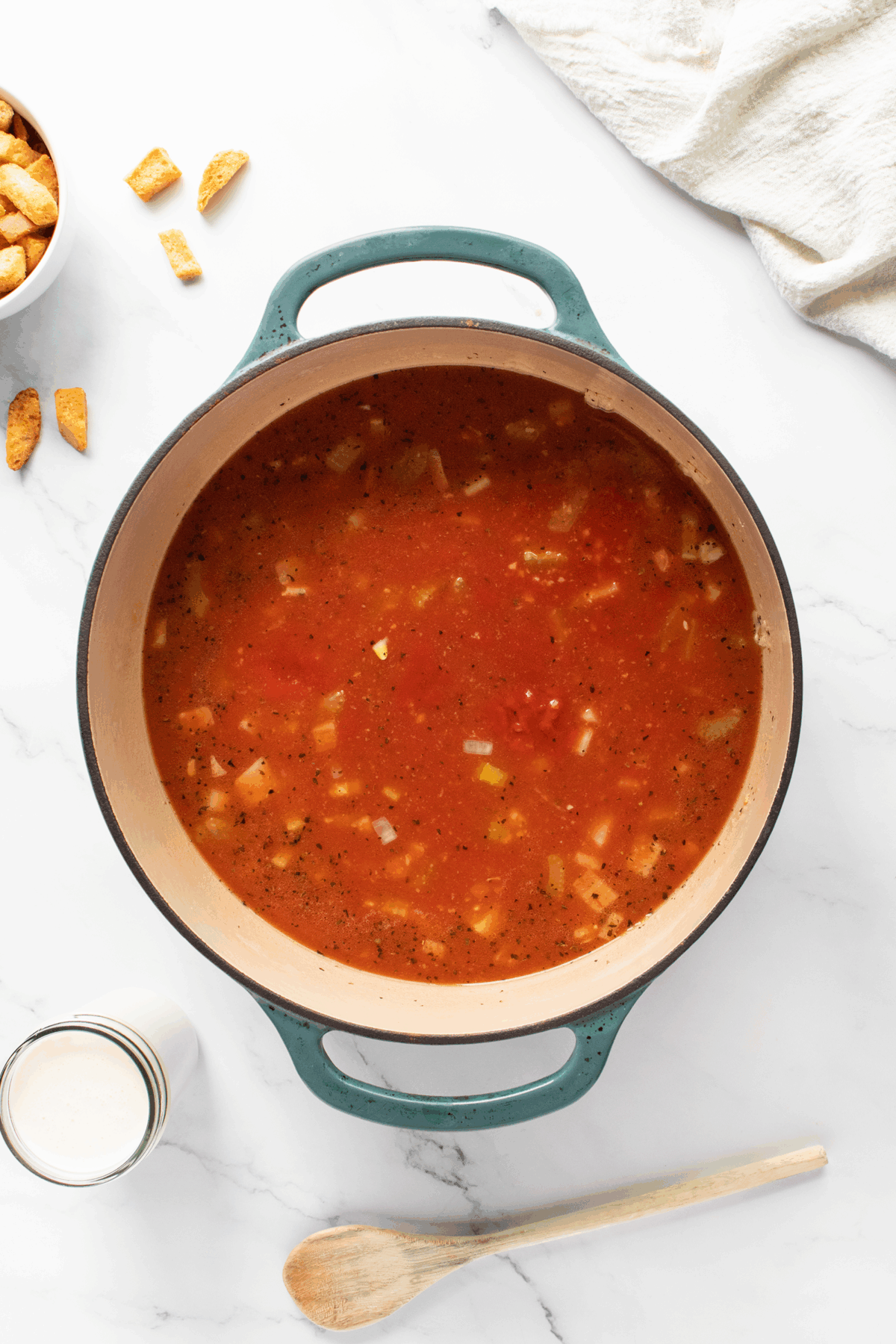 A large pot of tomato-based vegetable soup sits on a white marble surface, surrounded by a jar of cream, a bowl of croutons, a wooden spoon, and a white napkin.