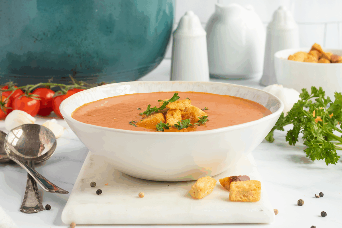 A bowl of creamy tomato soup topped with croutons and herbs sits on a white marble board, surrounded by fresh parsley, tomatoes, garlic, peppercorns, and a spoon. White dishes and a teal pot are in the background.