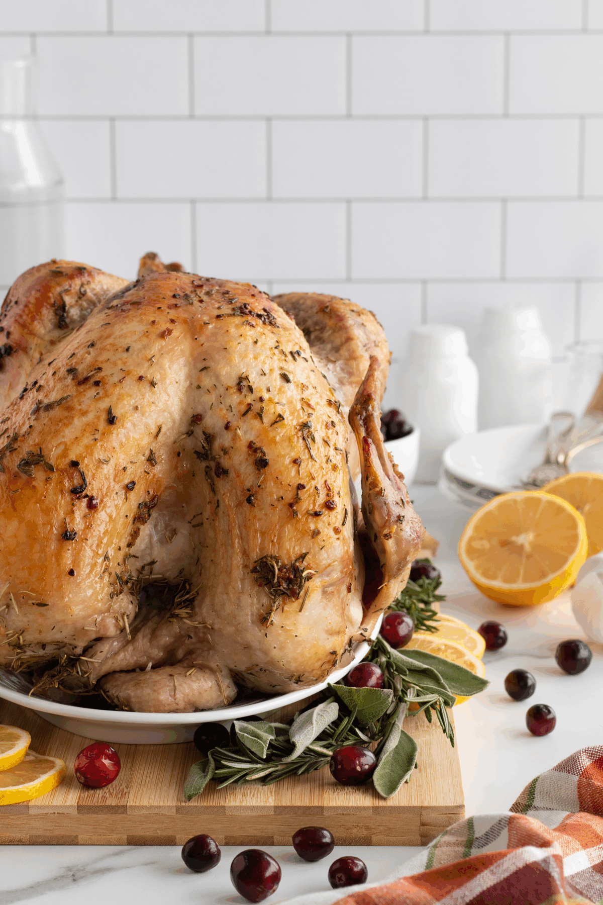 A roasted whole turkey is served on a white platter, garnished with fresh herbs, lemon slices, and cranberries. In the background are plates, utensils, and a white tile wall.