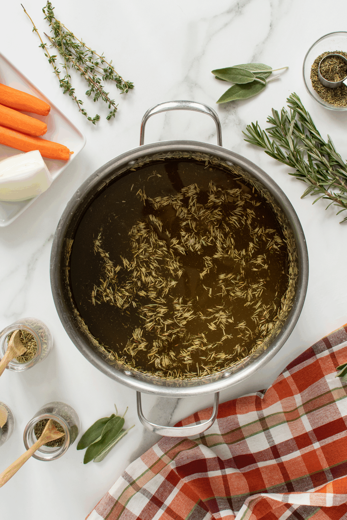 A pot of broth with floating herbs sits on a marble surface, surrounded by carrots, onion, fresh herbs, jars of spices, and a red plaid cloth.