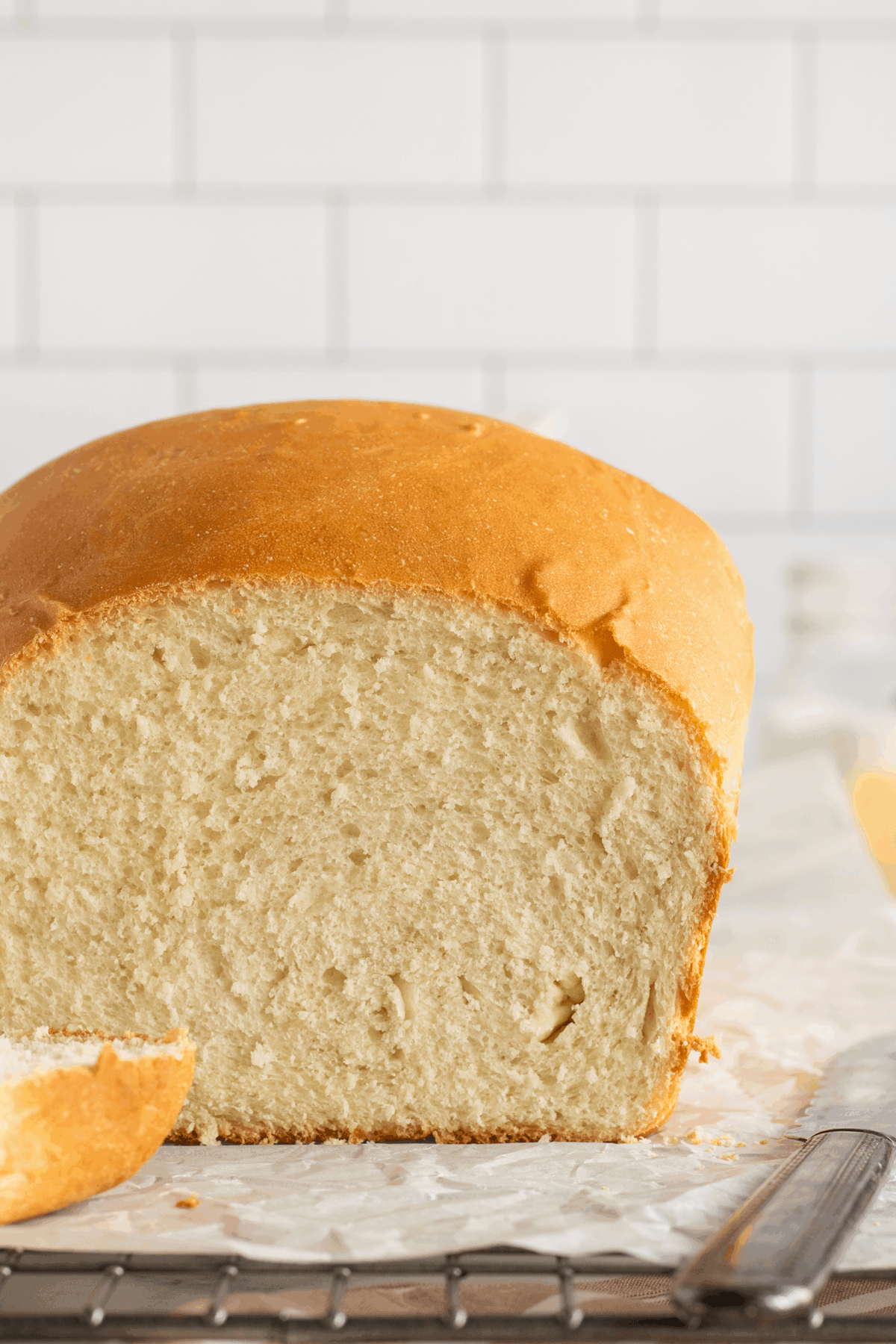 A loaf of homemade bread with a golden-brown crust sits on parchment paper; one slice is cut and lying nearby. White subway tiles are in the background.