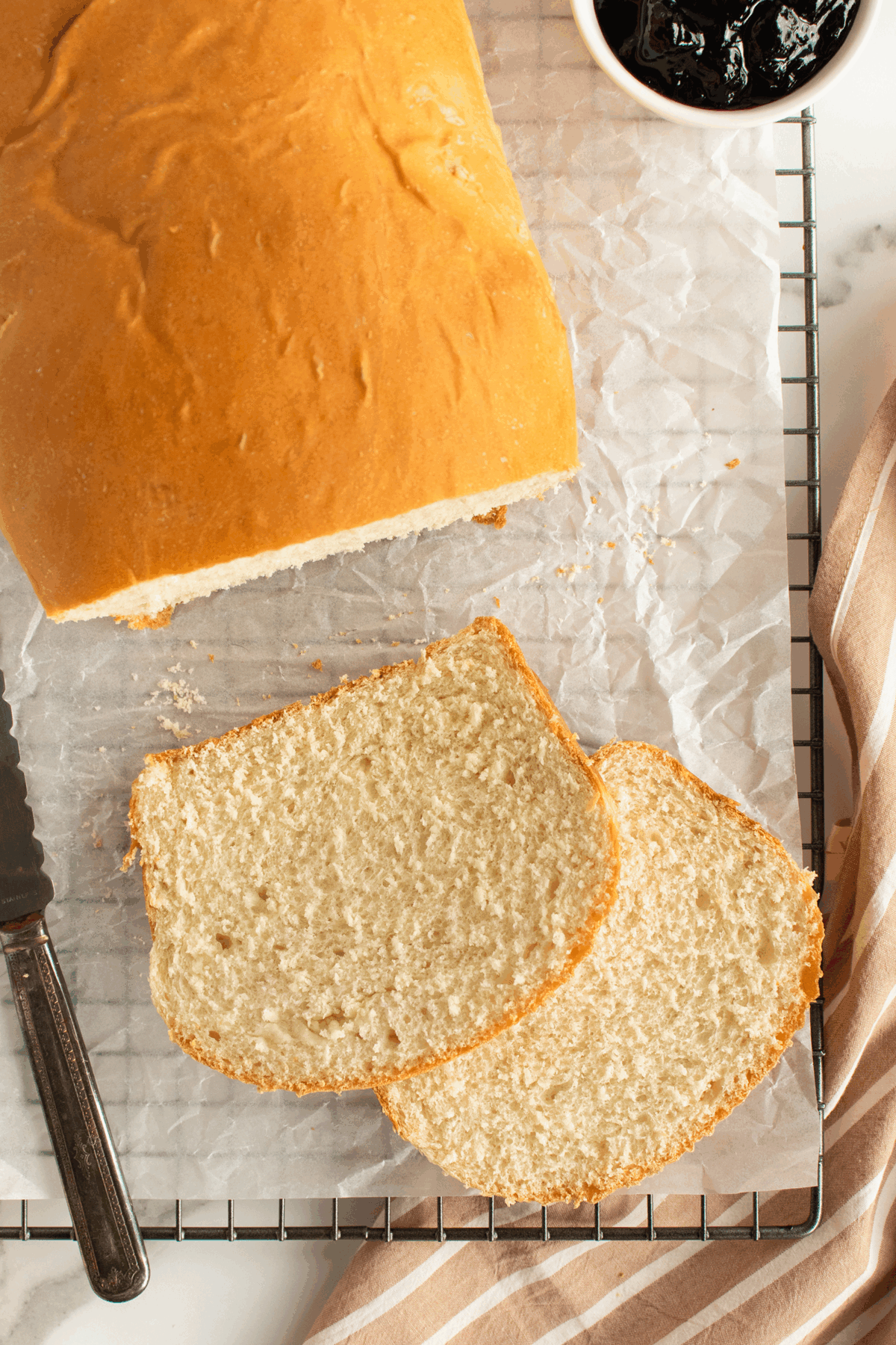 A loaf of bread with two slices cut, resting on parchment paper atop a cooling rack. A knife is nearby, and a small bowl of dark jam and a striped cloth are visible on the side.