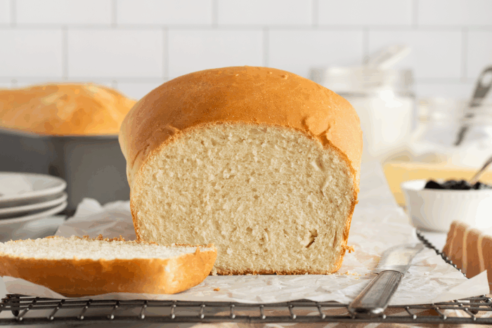 A loaf of homemade white bread sits on parchment paper with one slice cut. A bread knife rests nearby, and kitchen items are blurred in the background. The scene is bright and inviting.
