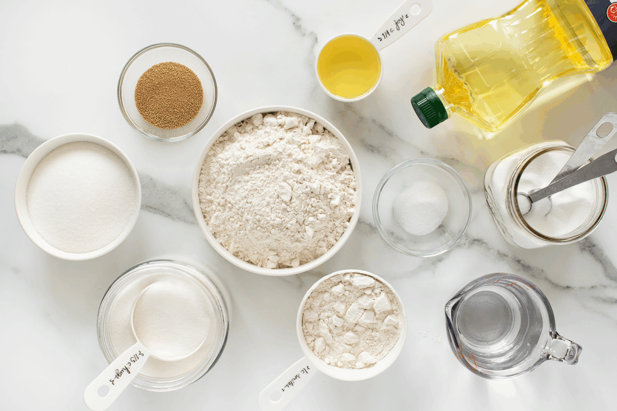 Various bread-making ingredients arranged on a white surface, including flour, sugar, yeast, salt, oil, water, and measuring cups, viewed from above.
