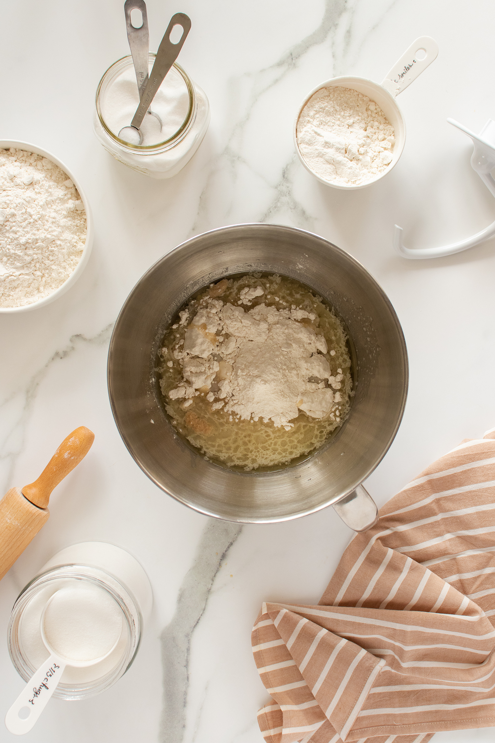 A metal mixing bowl with flour and wet ingredients sits on a marble countertop, surrounded by measuring cups with flour and sugar, a jar, a rolling pin, and a striped beige kitchen towel.