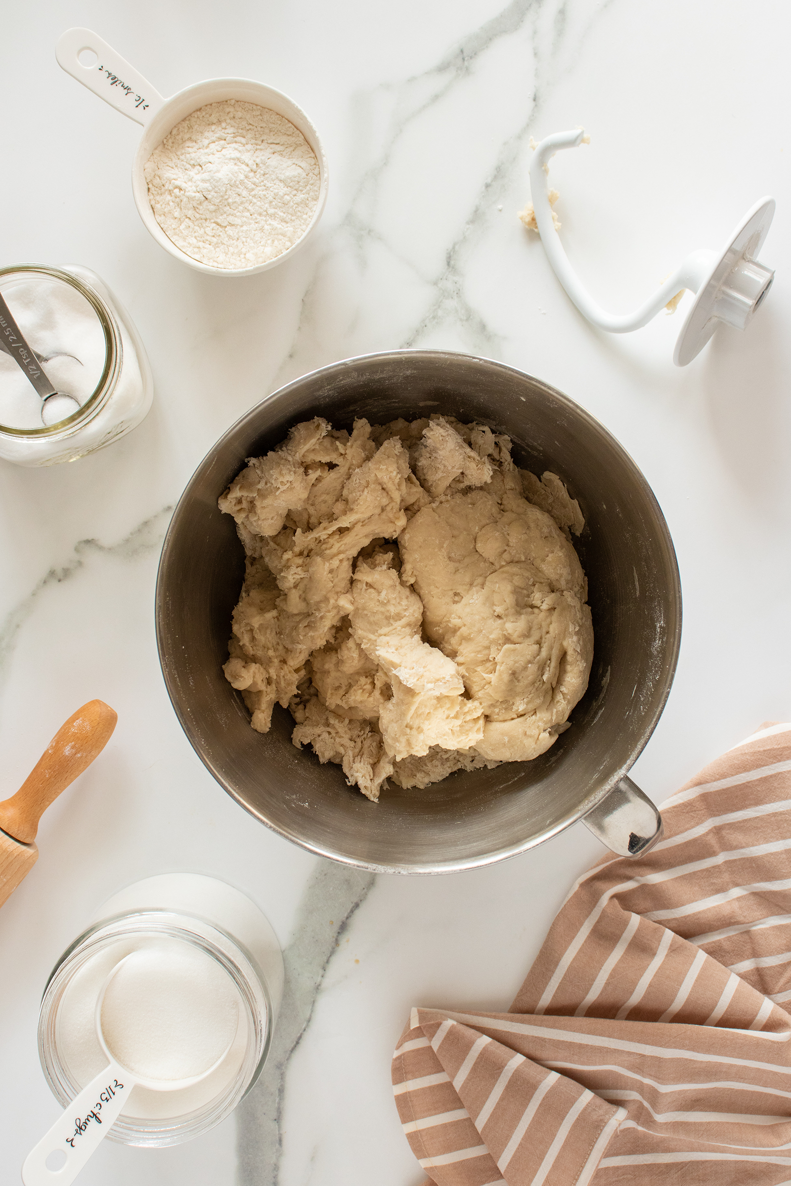 A metal mixing bowl filled with dough sits on a marble countertop, surrounded by measuring cups of flour and sugar, a jar of sugar, a dough hook, and a striped kitchen towel.