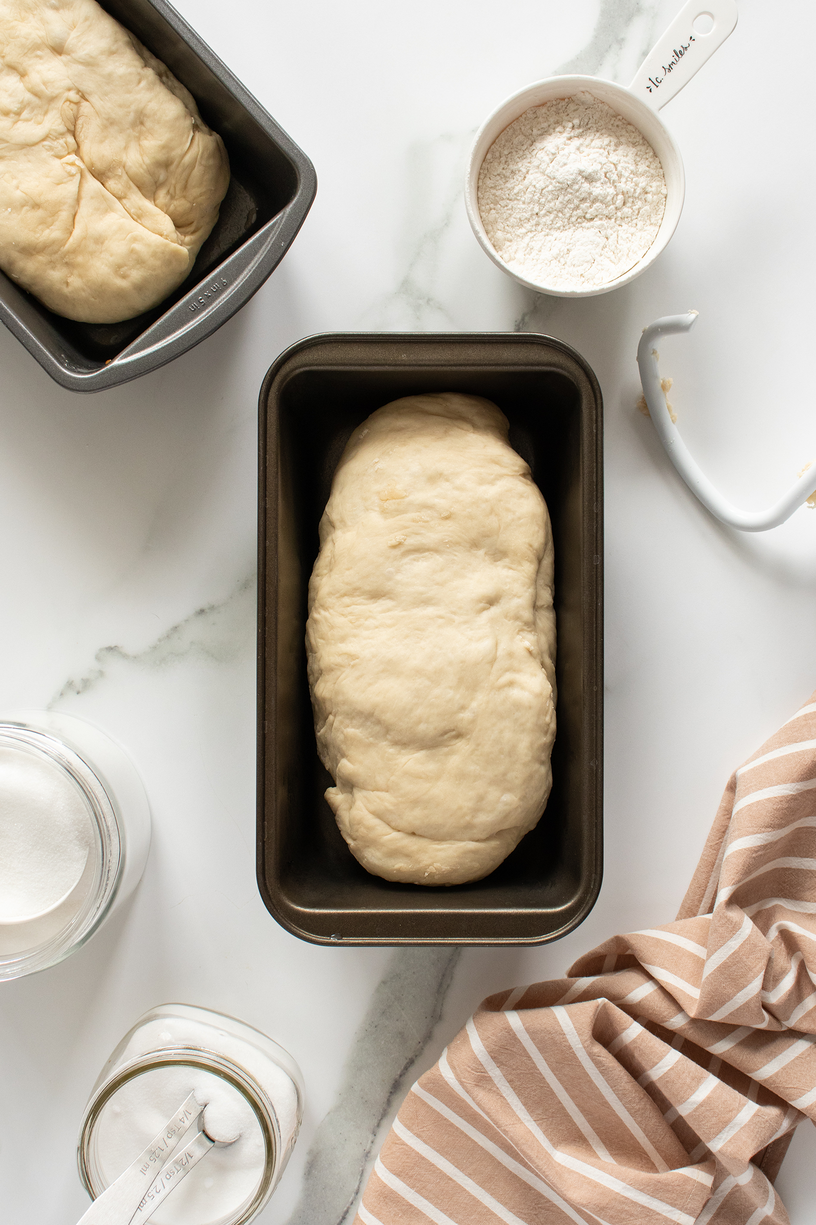 A loaf of raw bread dough sits in a rectangular baking pan on a marble surface, surrounded by a measuring cup with flour, a jar of sugar, a metal dough hook, and a brown-striped kitchen towel.
