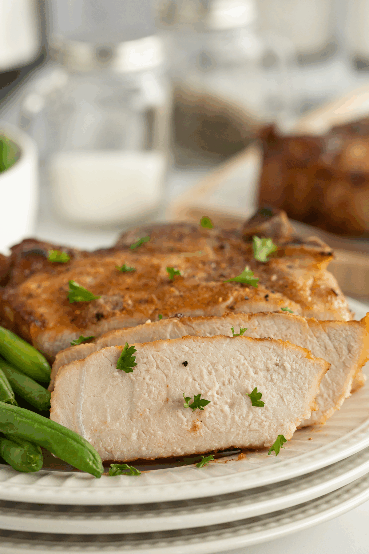 A close-up of a cooked pork chop, sliced to show its juicy interior, garnished with parsley and served with fresh green beans on a white plate. Glass jars are blurred in the background.