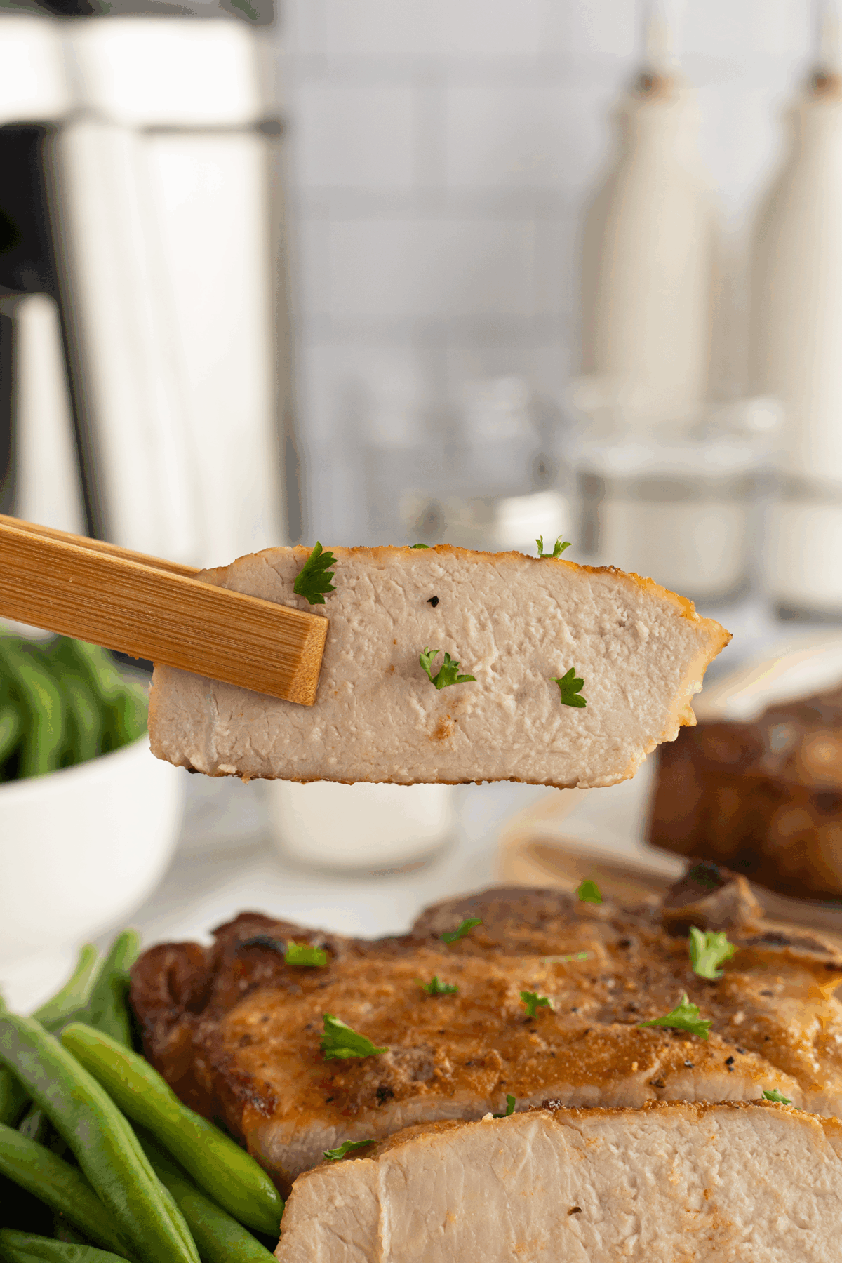 A close-up of a cooked pork chop held by chopsticks, garnished with parsley. More pork chops and green beans are visible on a plate in the background, with kitchen items blurred behind them.